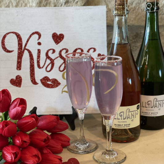 Two champagne flutes filled with Raider West Farm's recipe for Lavender French 75. Two bottles of Gallivant Bubbly Wine, red tulips, a white sign with the word “Kisses” and red hearts and lips on it are next to the Lavender French 75 filled glasses.