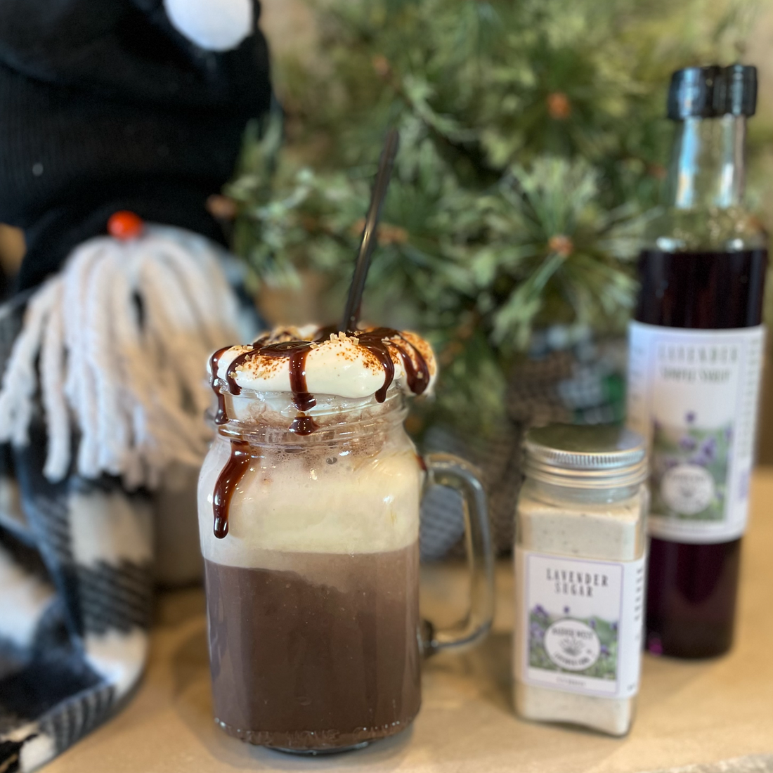 Glass mason jar filled with Dirty Snowman Lavender hot chocolate topped with toasted marshmallow and Raider West Farm's Lavender Hot Fudge. Raider West Farm's Lavender Sugar and Lavender Simple Syrup along with a winter gnome are in the background. 