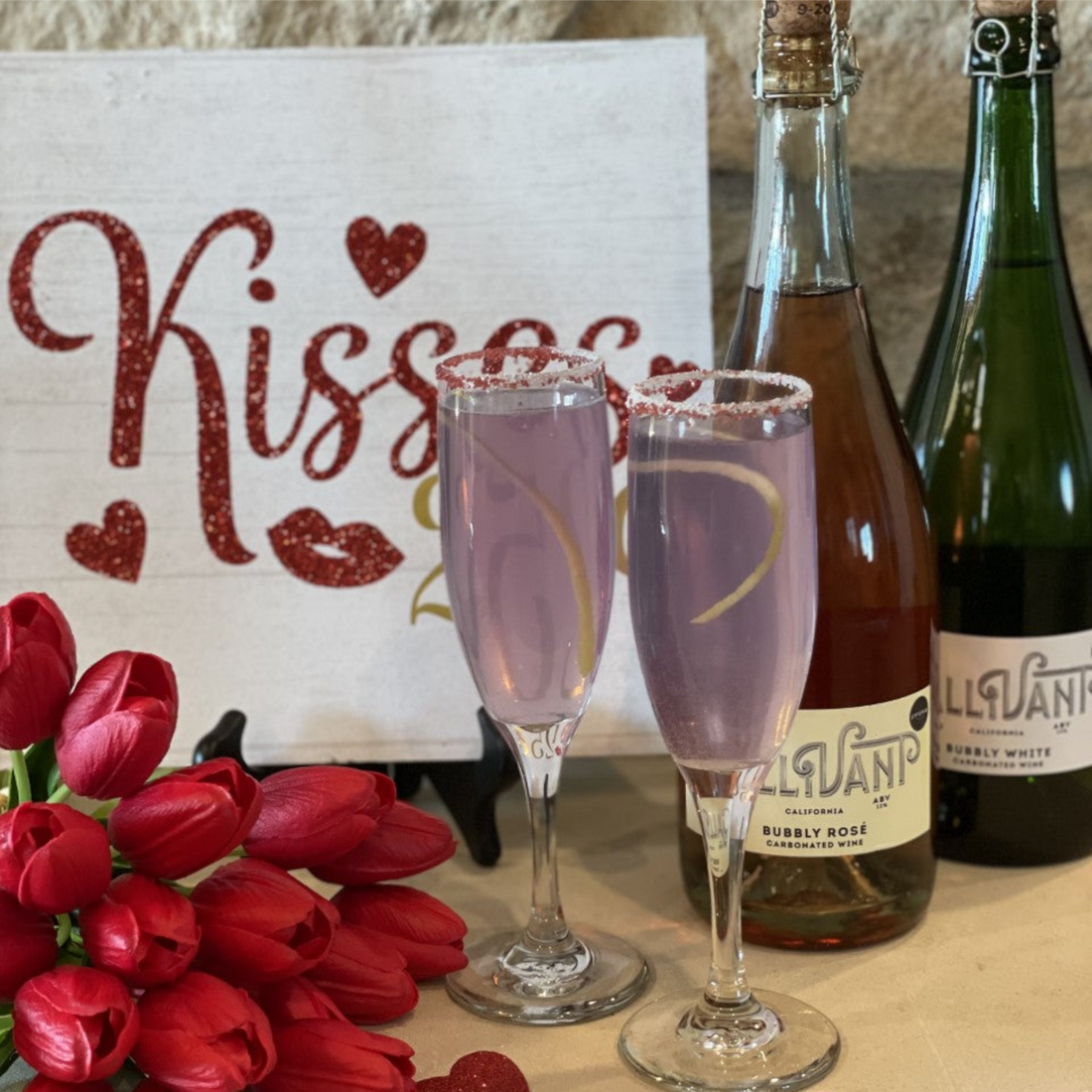 Two champagne flutes filled with Raider West Farm's recipe for Lavender French 75. Two bottles of Gallivant Bubbly Wine, red tulips, a white sign with the word “Kisses” and red hearts and lips on it are next to the Lavender French 75 filled glasses. 